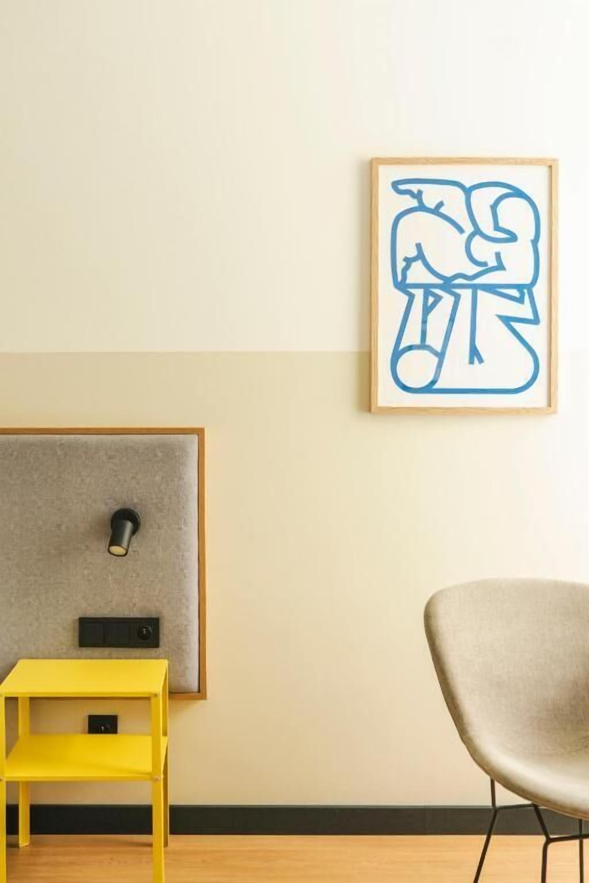 Una stanza con una testiera grigia, un comodino giallo, una sedia grigia e un quadro astratto appeso alla parete.