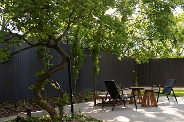 An outdoor patio with a table and chairs, surrounded by a dark fence and greenery.