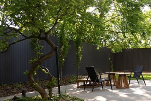 Um pátio externo com mesa e cadeiras, cercado por uma cerca escura e vegetação.