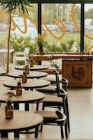 Restaurante com mesas e cadeiras redondas, plantas em vasos e uma grande janela com vista para carros estacionados e vegetação lá fora.