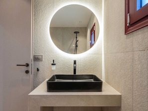 A bathroom with a round mirror, a rectangular sink, a towel dispenser, and a shower head.