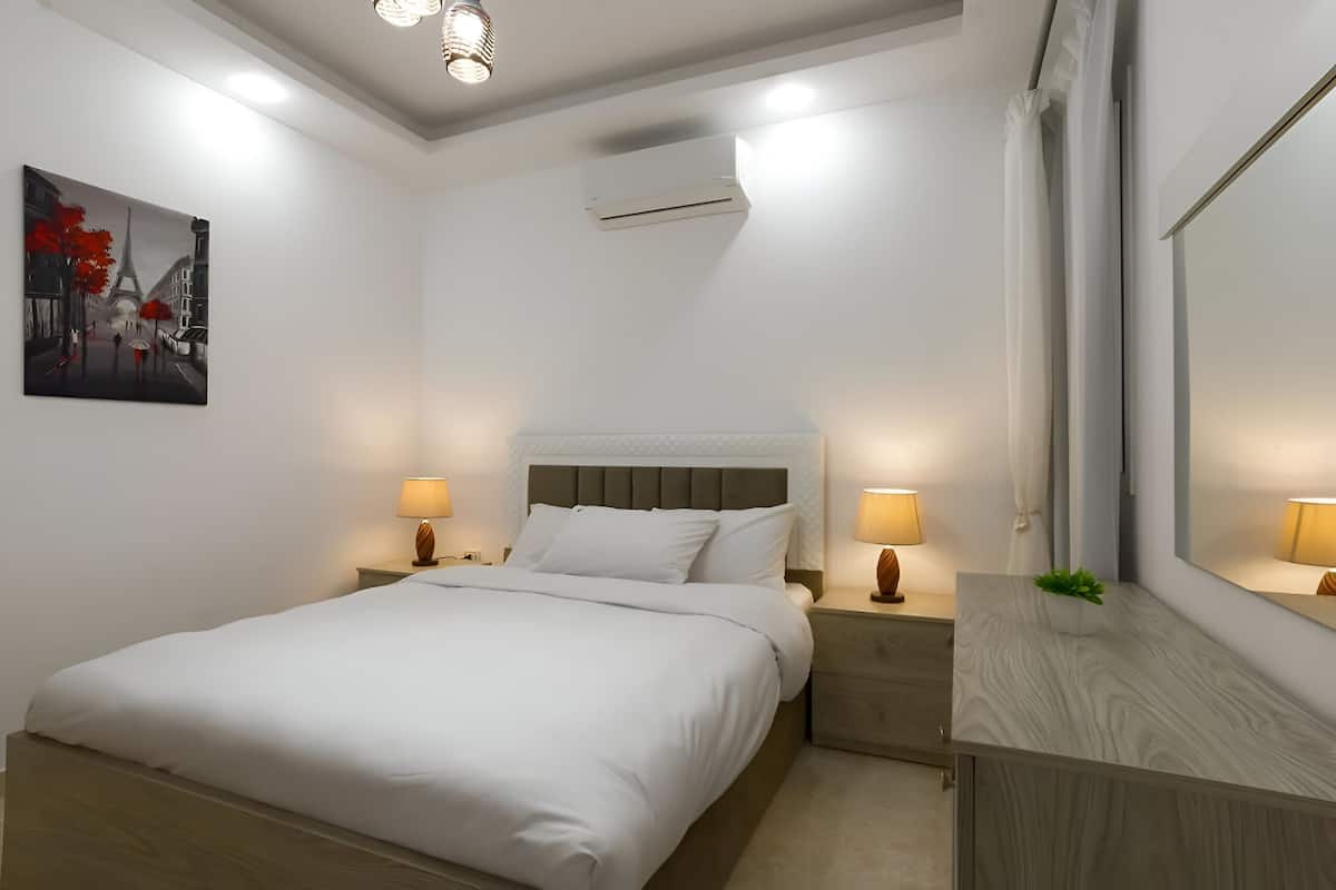 A neatly made bed with white linens, two bedside tables with lamps, a wall-mounted air conditioner, and a large mirror above a wooden dresser.