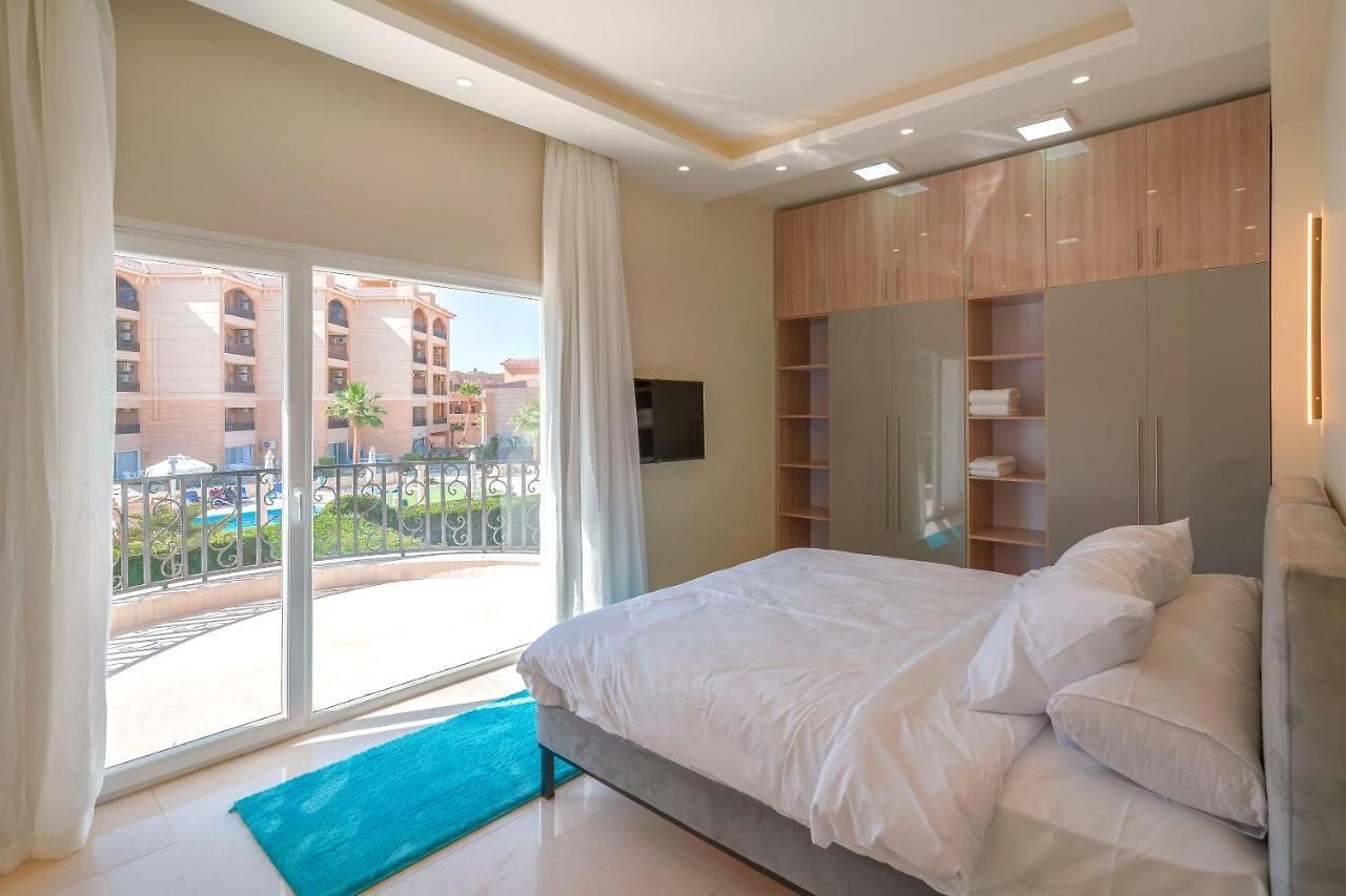 A modern hotel room with a large bed, a TV, and a balcony with a view of the pool and buildings.