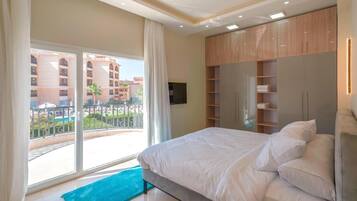 A modern hotel room with a large bed, a TV, and a balcony with a view of the pool and buildings.
