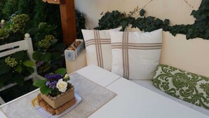Ein gemütlicher Sitzbereich im Freien mit einem weißen Tisch, einer gepolsterten Bank und einer dekorativen Pflanzengestaltung.
