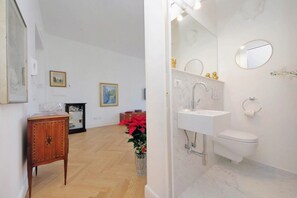 Un baño moderno con un lavabo blanco, espejo e inodoro. Hay un mueble de madera y un florero con flores rojas en el suelo.