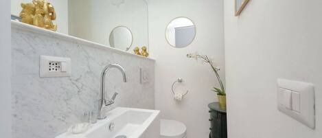 Une salle de bain avec un lavabo blanc, des toilettes et un miroir. On y trouve une statue dorée posée sur le comptoir, une plante en pot et un tableau encadré accroché au mur.