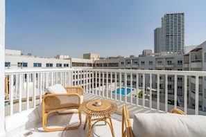 A balcony with wicker furniture, a pool, and high-rise buildings in the background. - Beautiful Studio in JVC near Halfa Park (Dubai)