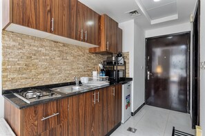 A kitchen with wooden cabinets, a stone wall backsplash, a black countertop, a stainless steel sink, and a coffee machine. - Beautiful Studio in JVC near Halfa Park (Dubai)