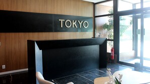 En receptionsdisk med en skylt som visar texten ”TOKYO” i en modern hotellobby med en väggpanel i trä, en glasdörr och en liten statyett på bordet.