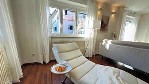 A modern hotel room with a large window, a sofa, a small table with a croissant, and a lamp. - Scharnhäusle 2- Large apartment with bathtub & balcony (Ostfildern)
