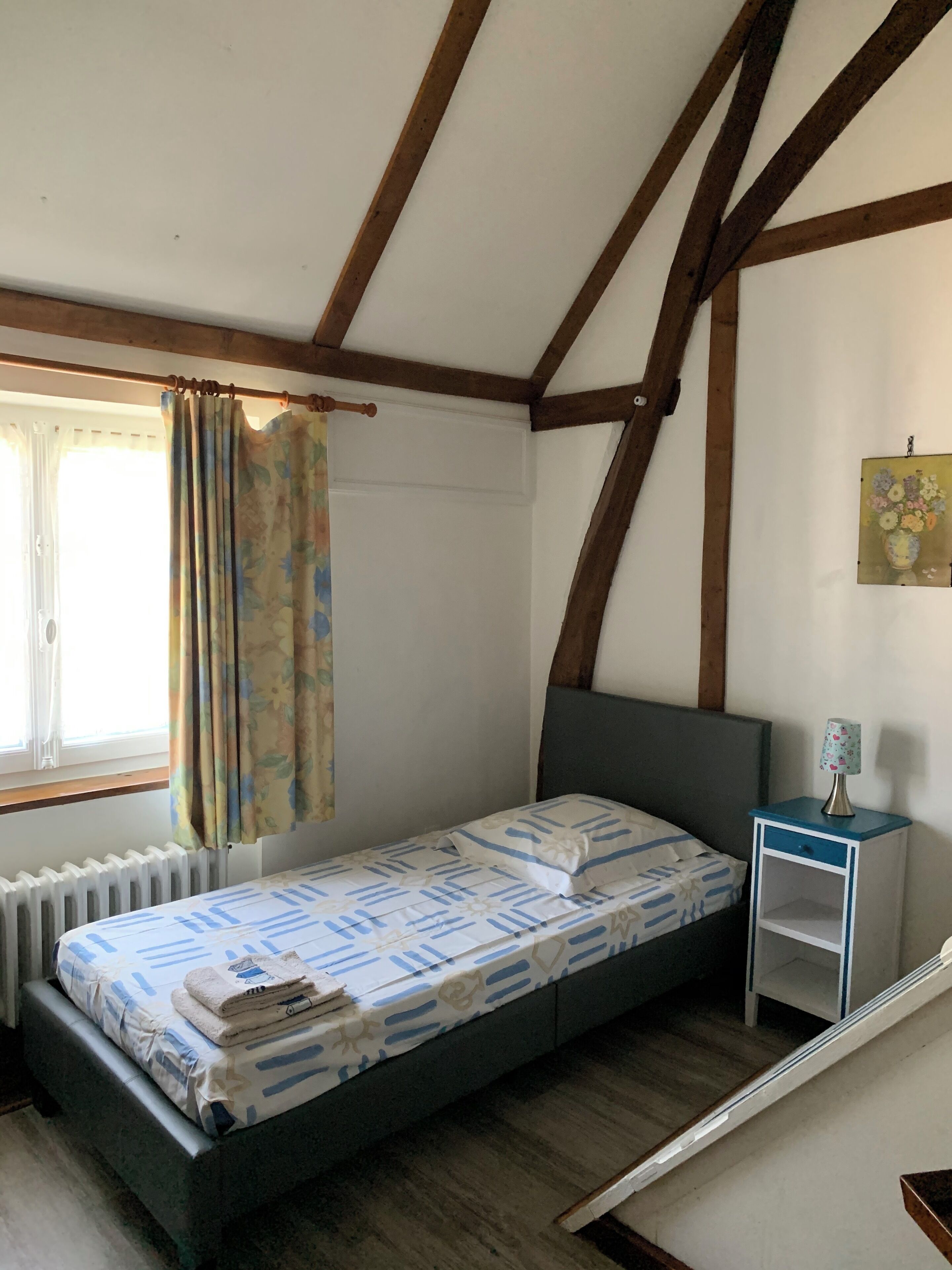 Ein Einzelbett mit einem gemusterten Überwurf, ein Nachttisch mit Lampe, ein Heizkörper und ein Fenster mit Vorhängen.