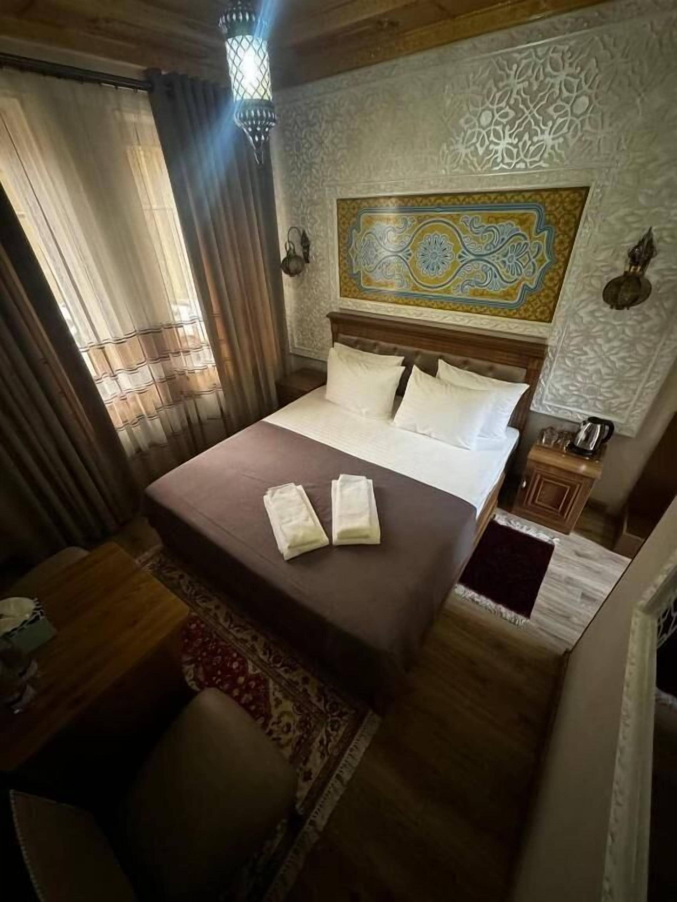 A hotel room with a large bed, a wooden headboard, a decorative wall hanging, and a small nightstand with a lamp.