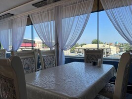 A hotel room with a large bed, a table, and chairs. There are large windows with sheer curtains.