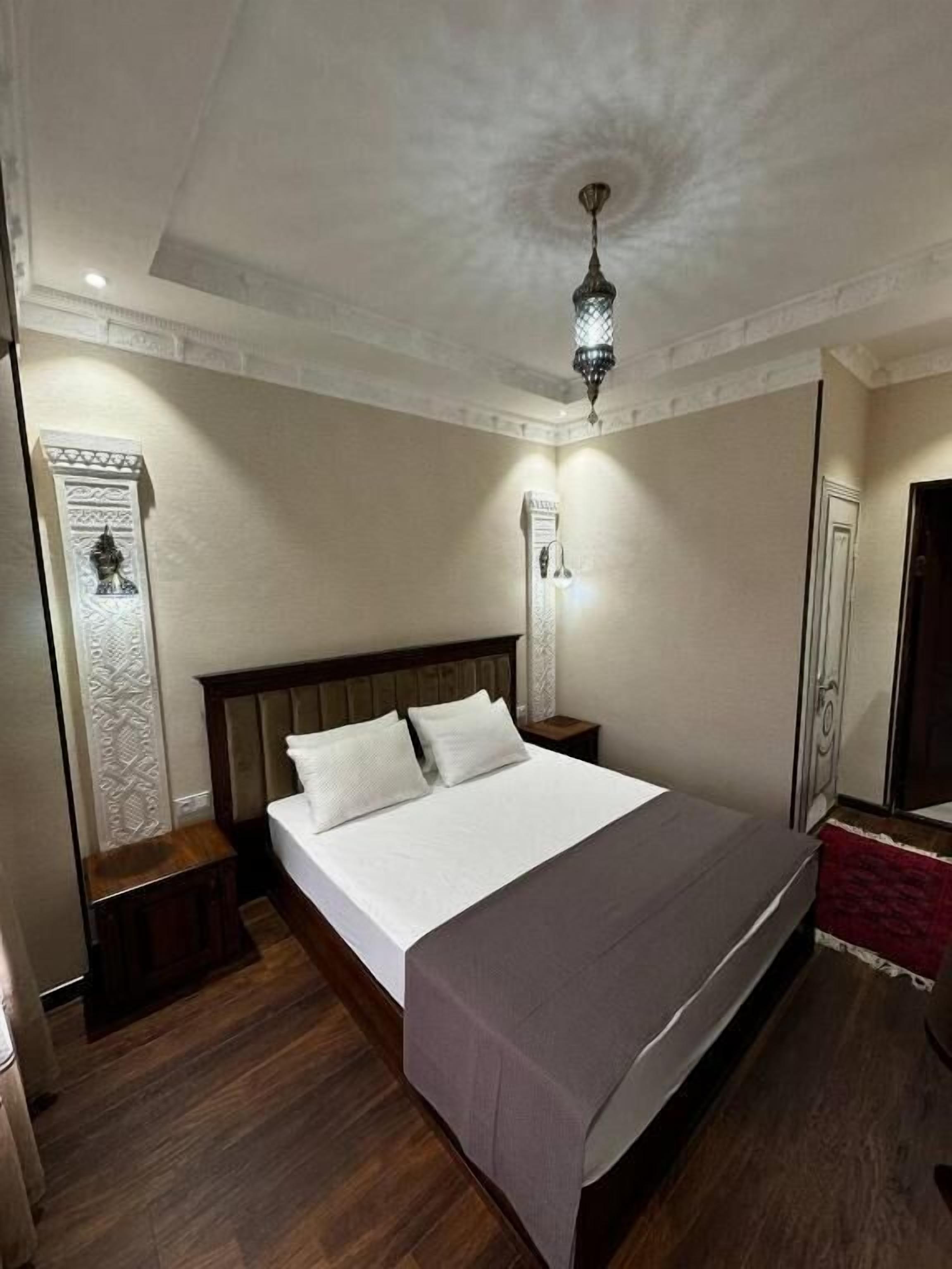 A hotel room with a large bed, wooden headboard, a nightstand, a wall-mounted light, and a decorative wall panel.