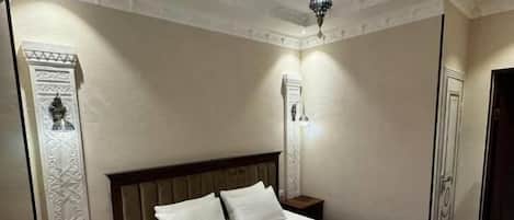 A hotel room with a large bed, wooden headboard, a nightstand, a wall-mounted light, and a decorative wall panel.