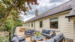 Un espace patio aménagé avec du mobilier d’extérieur, comprenant un canapé, des fauteuils et une table, entouré d’un jardin et d’un bâtiment doté de fenêtres.