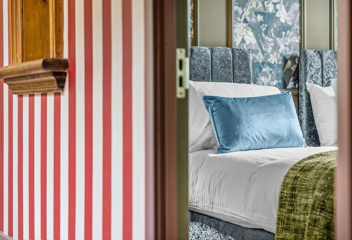 Ein Bett mit blauem Kissen und weißer Bettwäsche, eine gestreifte Wand und ein Fenster mit blumigen Vorhängen.