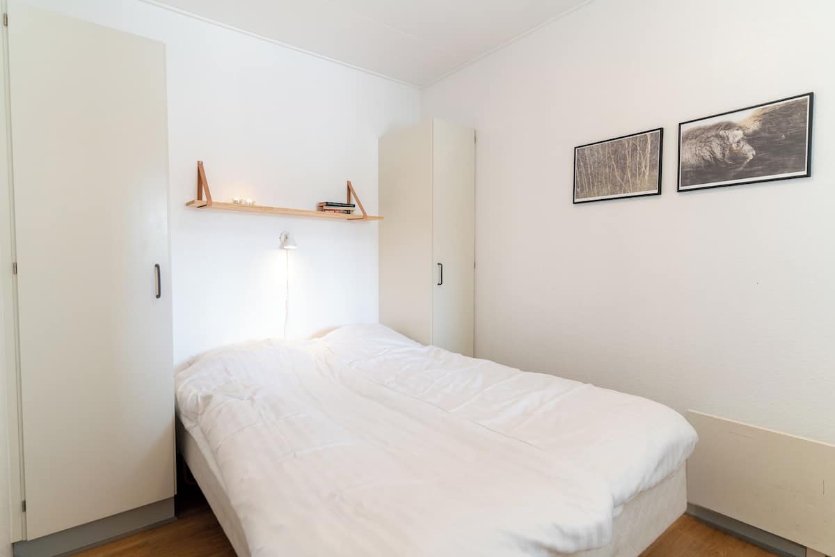 A bedroom with a bed, a wardrobe, a shelf with books, and two framed pictures on the wall.