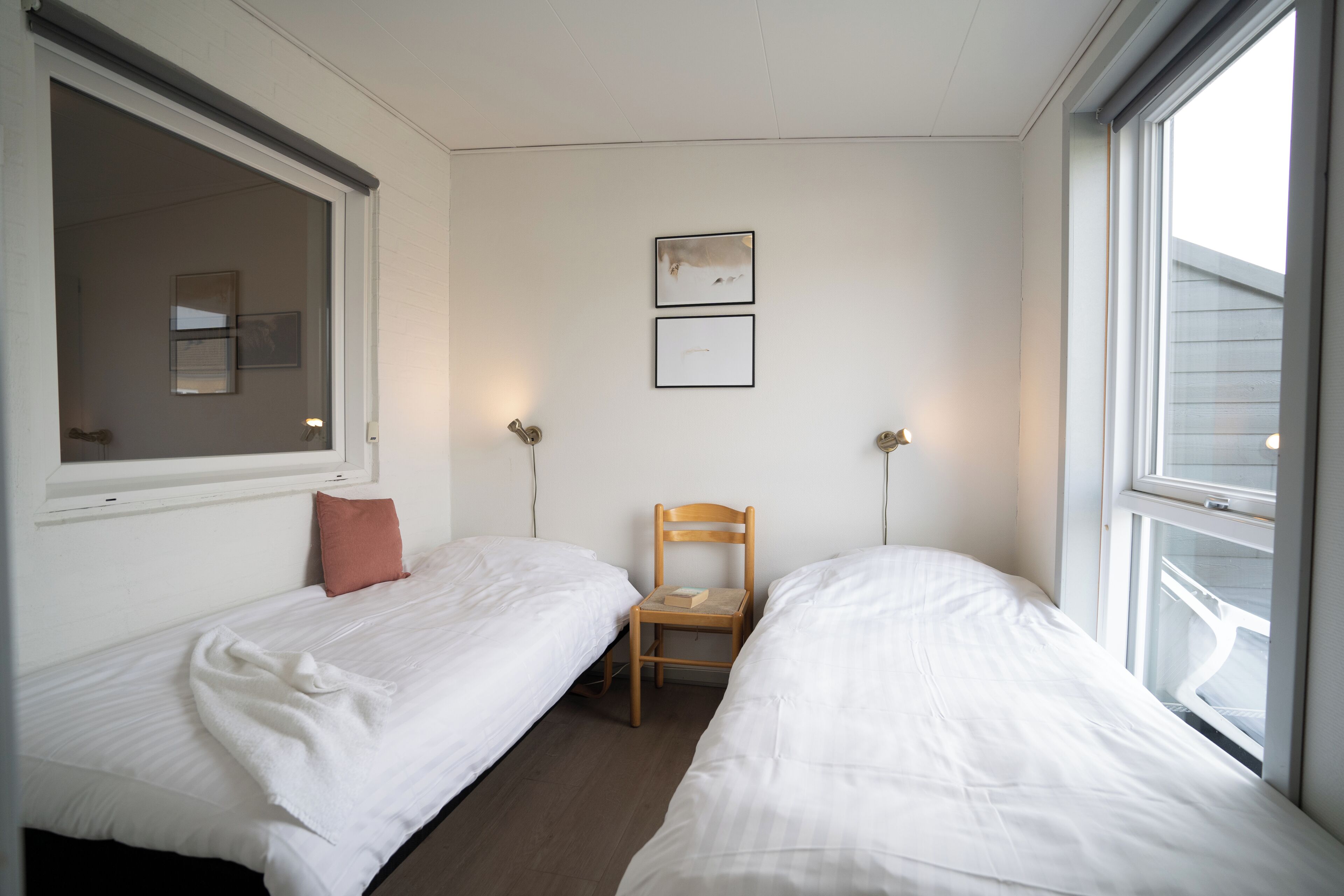 A room with two single beds, a wooden chair, a window, and two wall-mounted lamps.