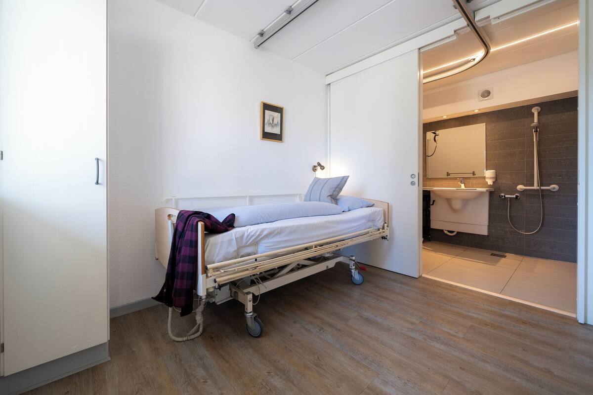 Une chambre d’hôpital avec un lit, une table de chevet, une petite fenêtre et une salle de bain équipée d’une douche et d’un lavabo.