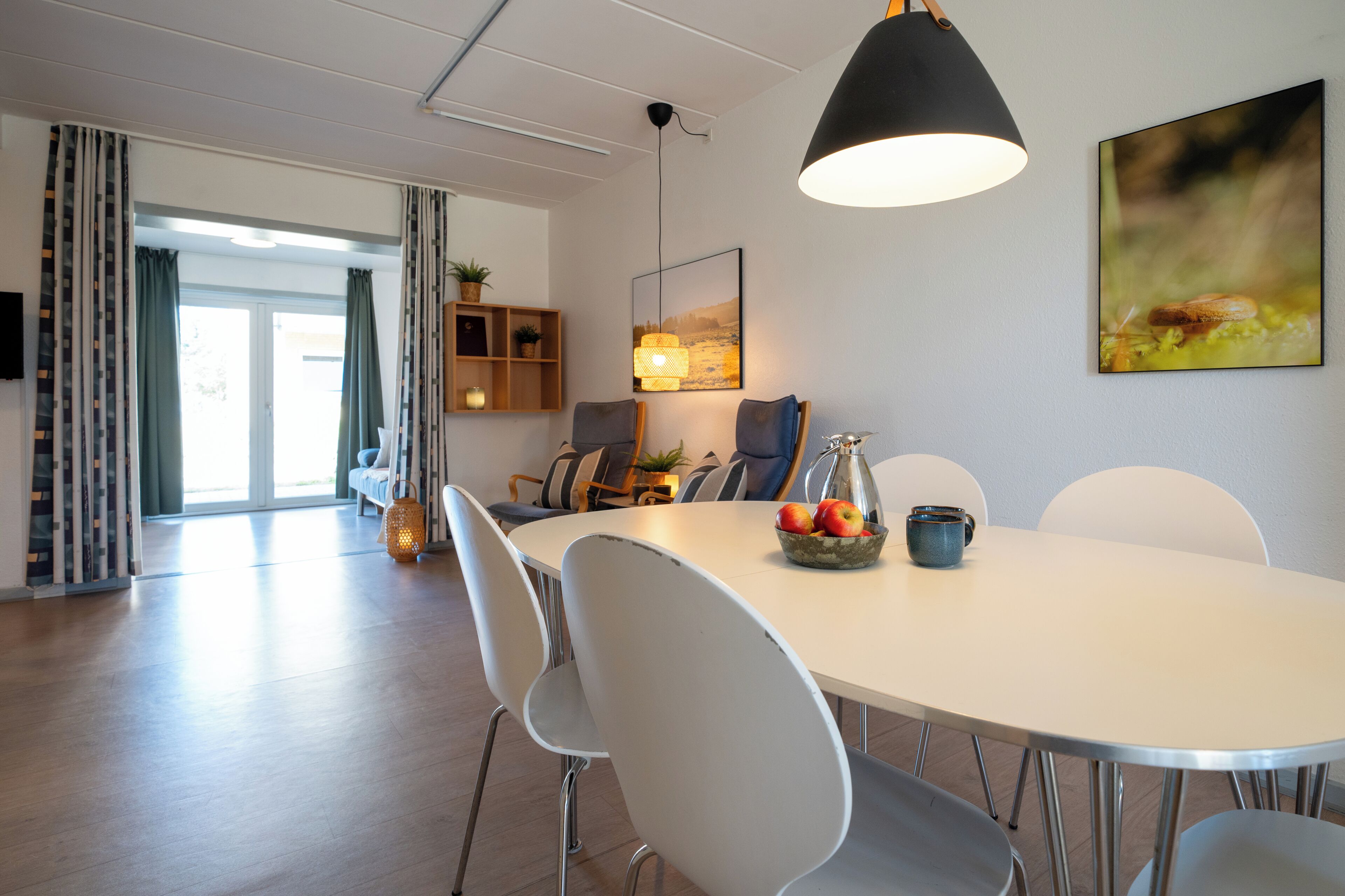 Ein moderner Essbereich mit einem runden Tisch, weißen Stühlen, einer schwarzen Pendelleuchte und einem gerahmten Bild an der Wand.