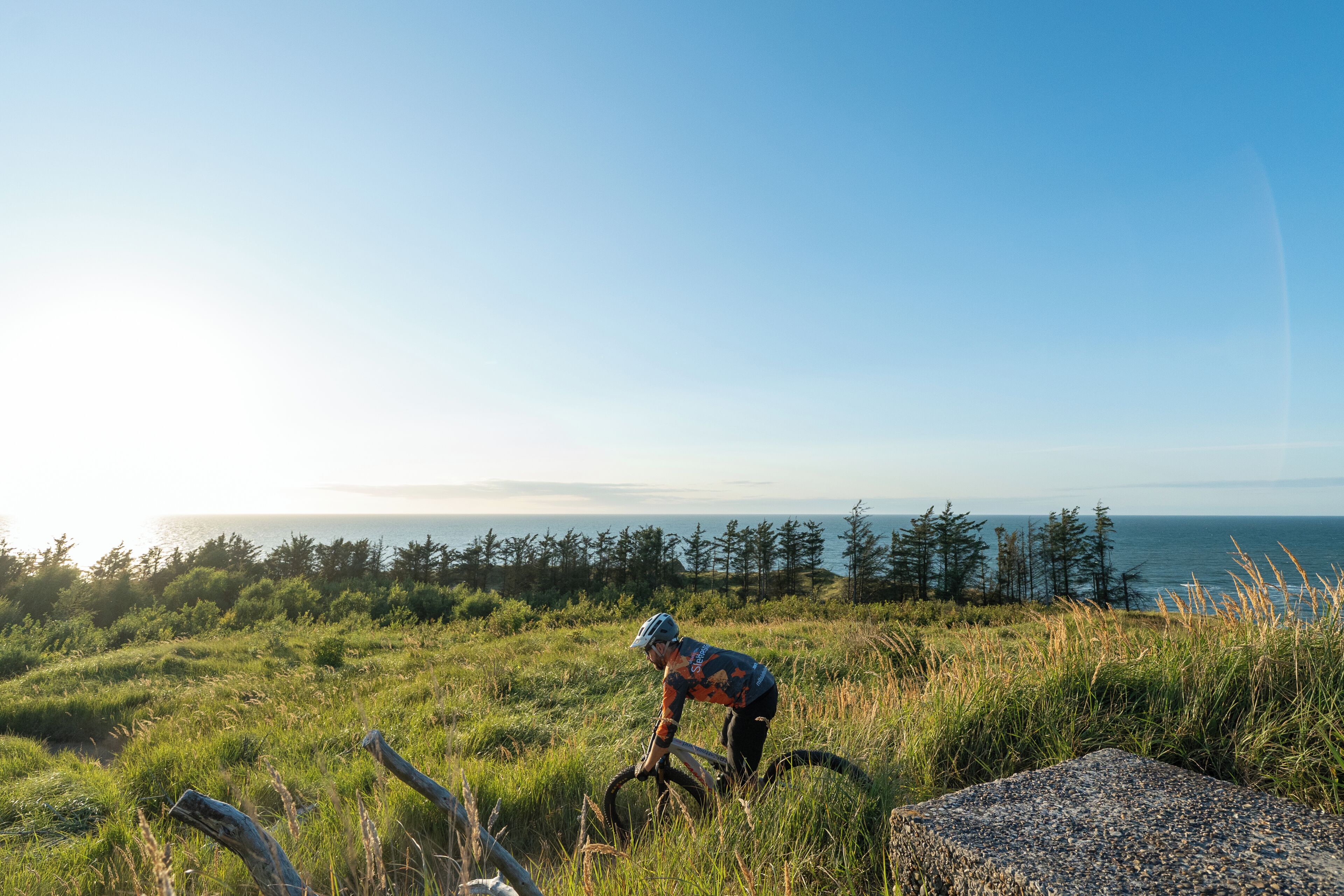 A person riding a mountain bike on a grassy field with a view of the ocean and trees in the background.