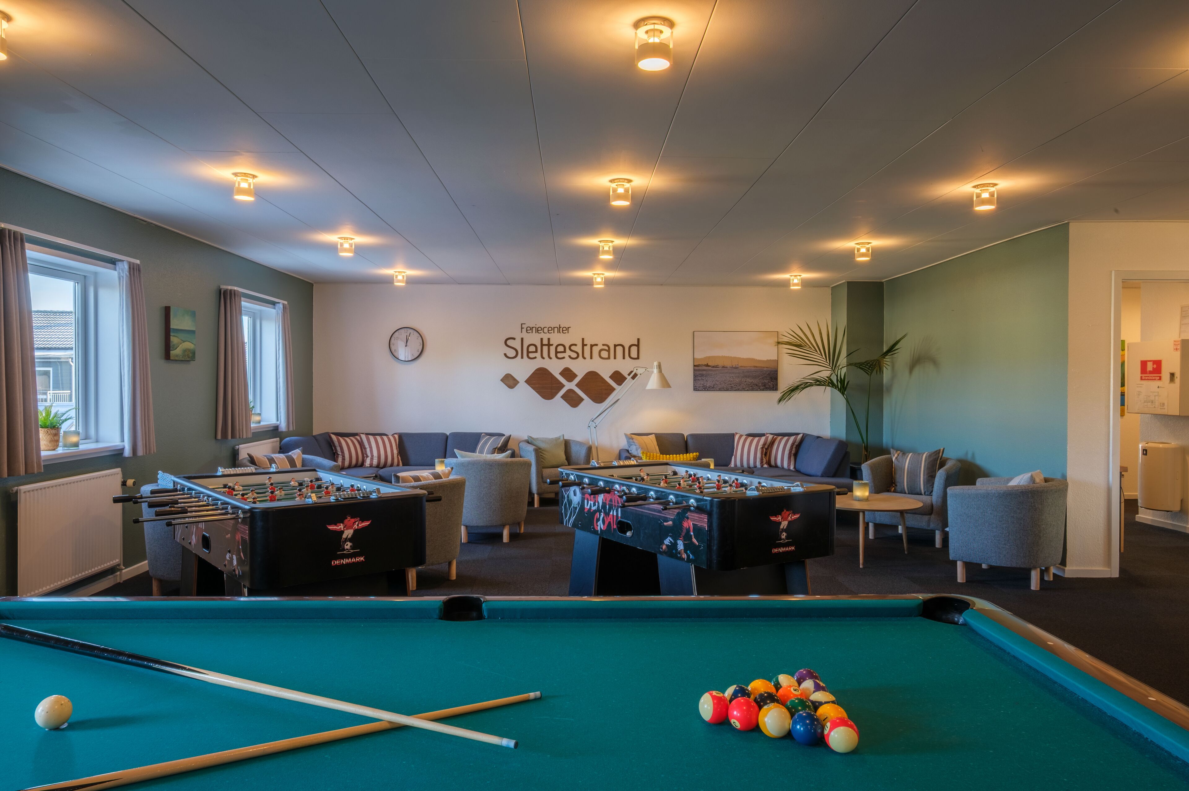 Ett biljardbord, två fotbollsspel, en soffa och en vägg med en klocka och en skylt med texten ”Fiencenter Slettesstrand”.