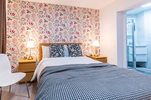 A hotel room with a floral wallpaper, a bed with a checkered bedspread, two bedside lamps, a white chair, and a bathroom visible through an open door. - Fulham 6BR 6BA Tube Family London (London)