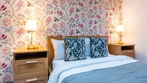 A bedroom with floral wallpaper, a wooden headboard, two bedside tables with lamps, and a bed with patterned pillows. - Fulham 6BR 6BA Tube Family London (London)