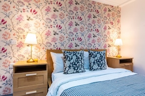 A bedroom with floral wallpaper, a wooden headboard, two bedside tables with lamps, and a bed with patterned pillows. - Fulham 6BR 6BA Tube Family London (London)