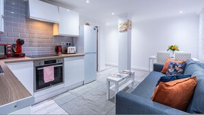 A modern kitchen with white cabinets, a built-in oven, and a refrigerator. There is a small dining area with a white table and a sofa with cushions. - Fulham 6BR 6BA Tube Family London (London)