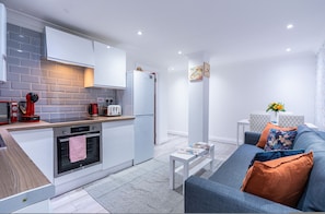 A modern kitchen with white cabinets, a built-in oven, and a refrigerator. There is a small dining area with a white table and a sofa with cushions. - Fulham 6BR 6BA Tube Family London (London)