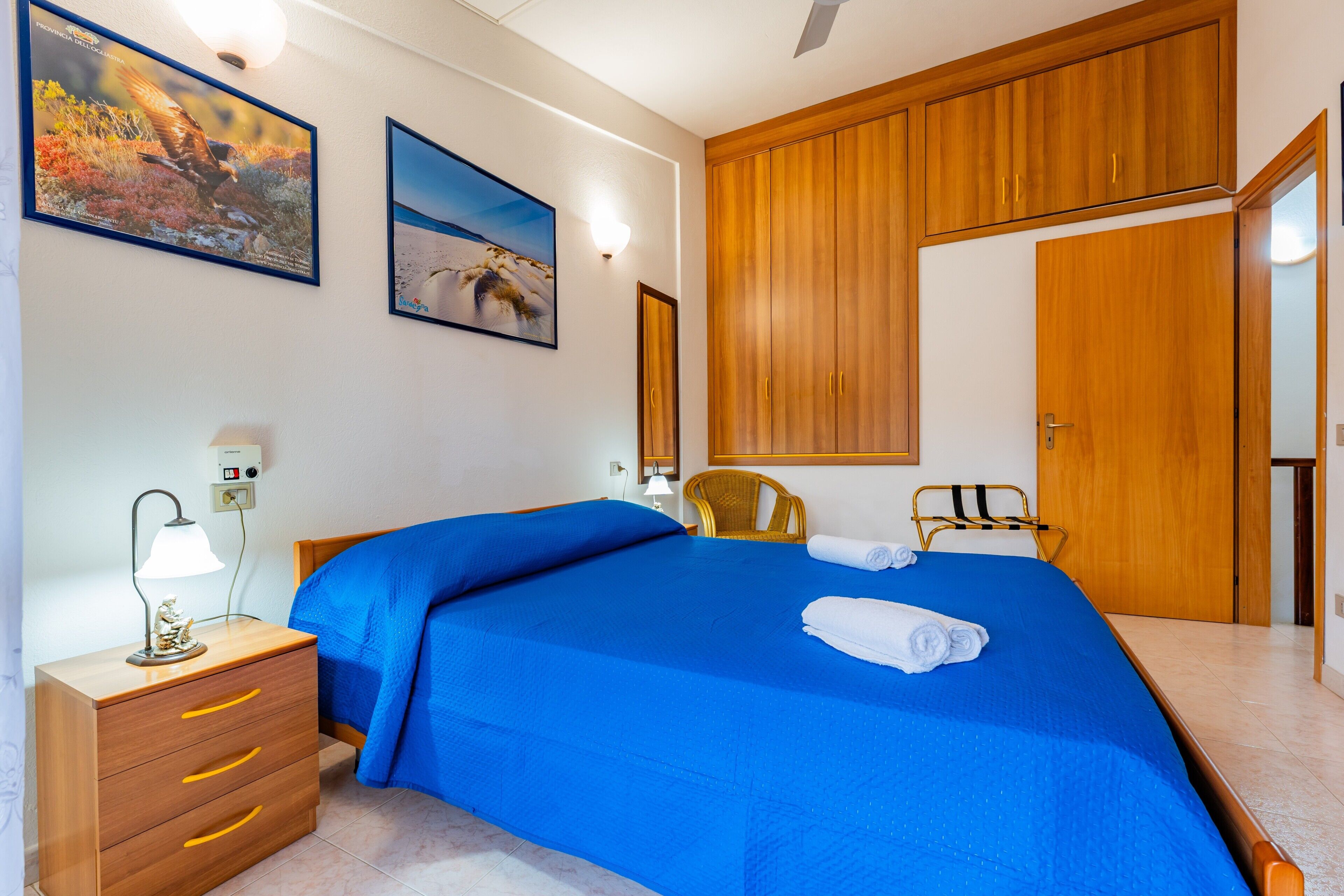 A bedroom with a bed, wooden wardrobe, two wall-mounted paintings, a bedside lamp, and a chair.