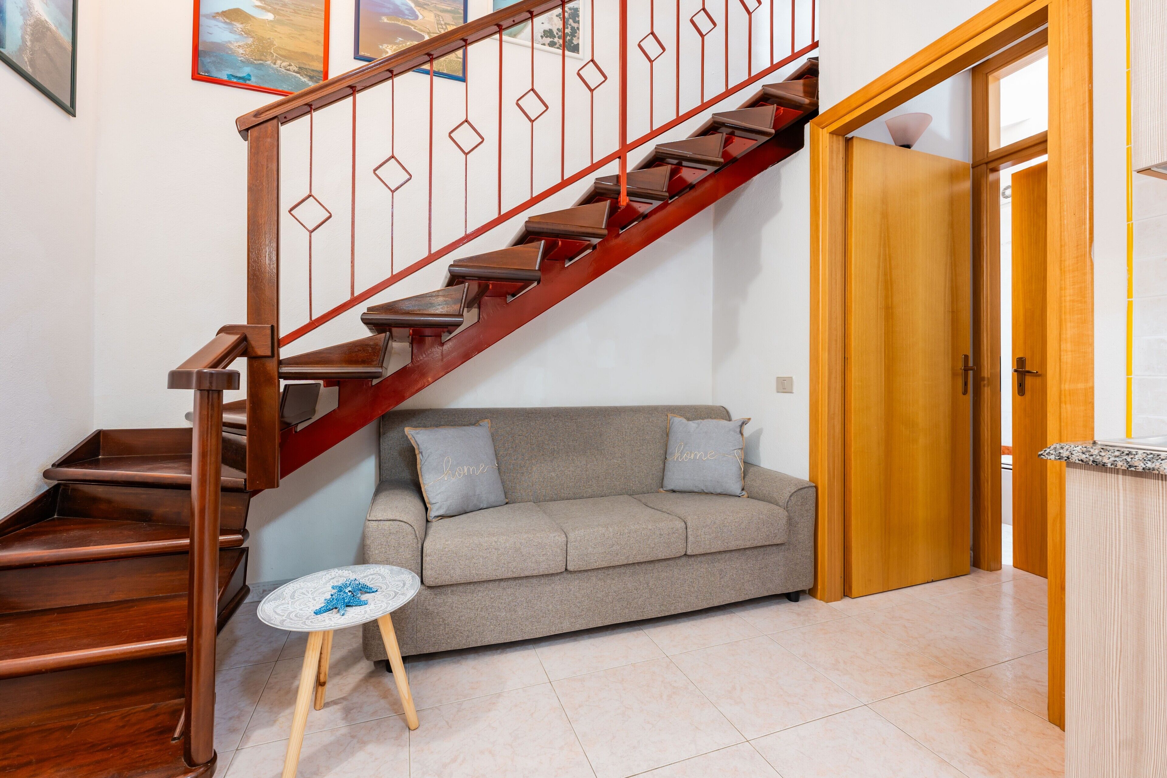 A staircase with wooden steps and a red railing, a grey sofa with two pillows, a small round table, and a wooden door.