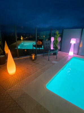Un espace extérieur avec une piscine, comprenant une chaise et une table, entouré de murs en verre et éclairé par des luminaires modernes.