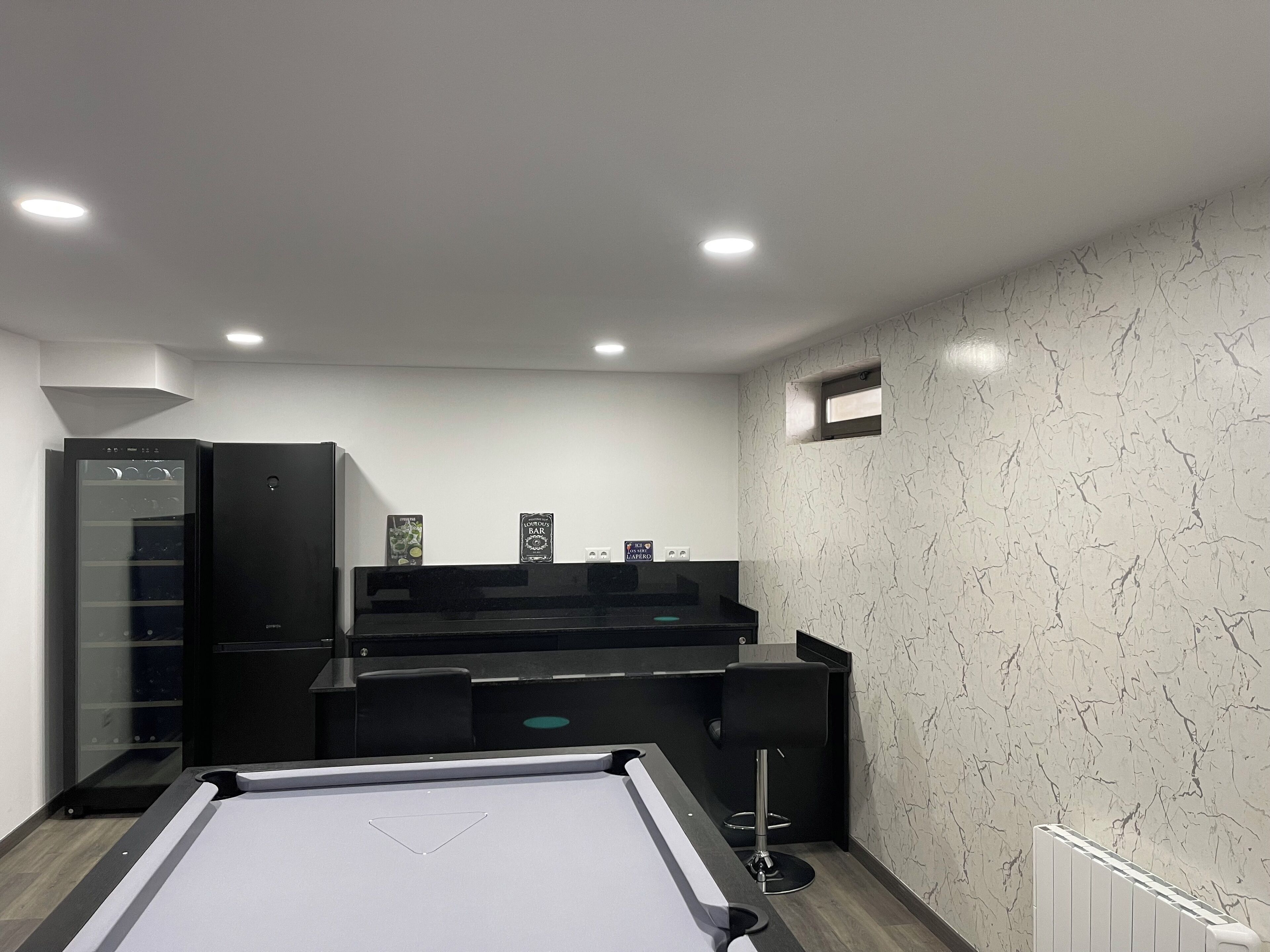 A pool table with a billiards cue, a bar area with stools, and a refrigerator.