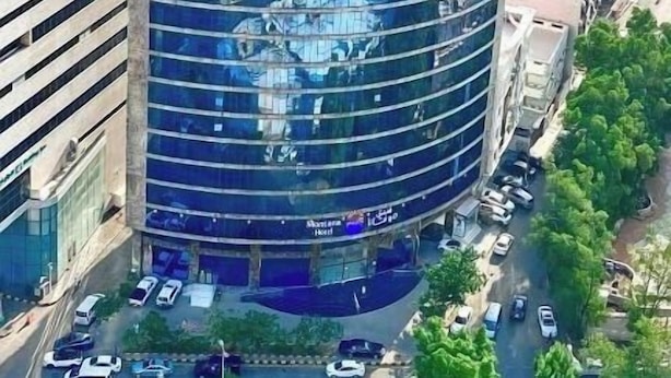 Un hotel moderno con fachada de vidrio curva, rodeado de edificios urbanos y un telón de fondo montañoso.