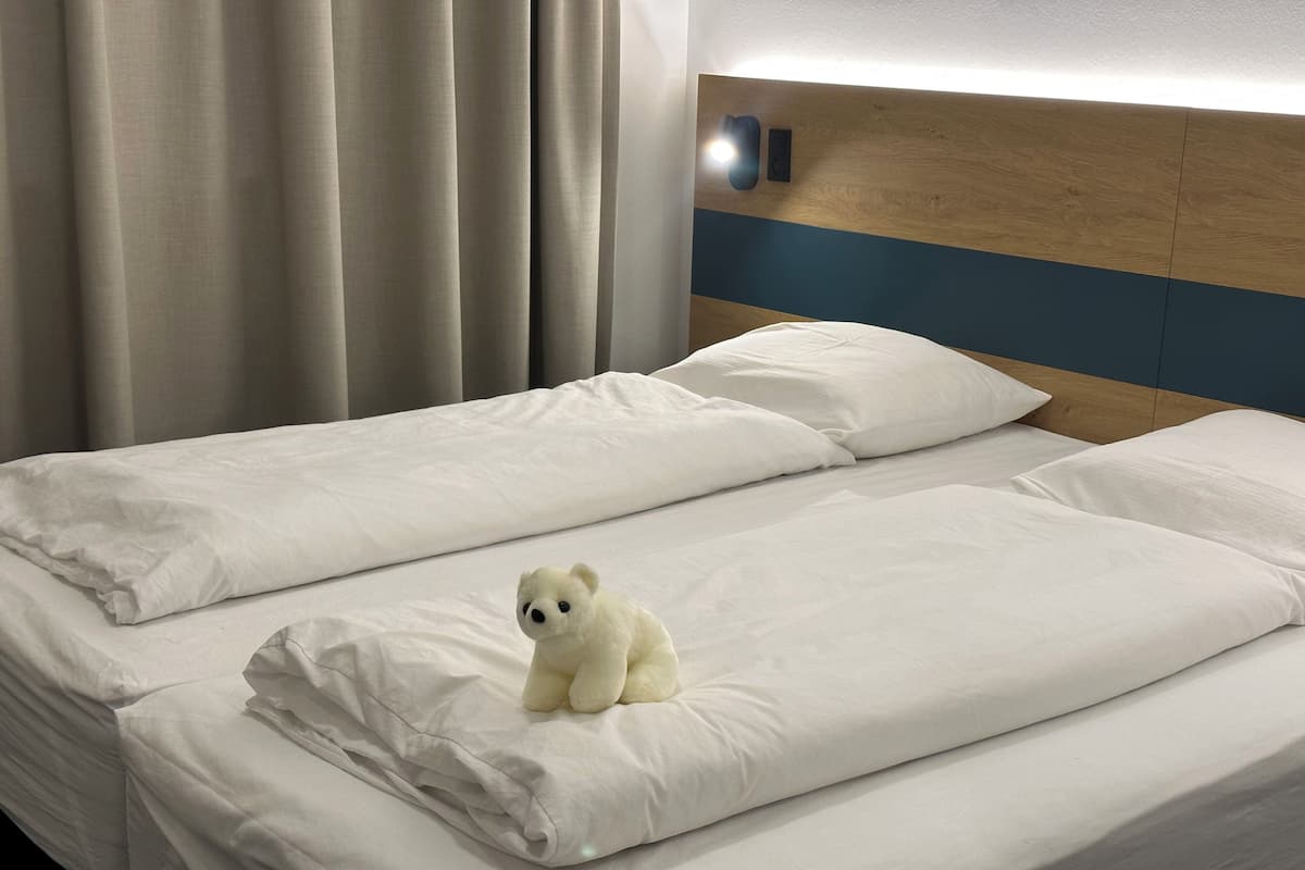 Una cama doble con ropa de cama blanca y un peluche pequeño a la izquierda.