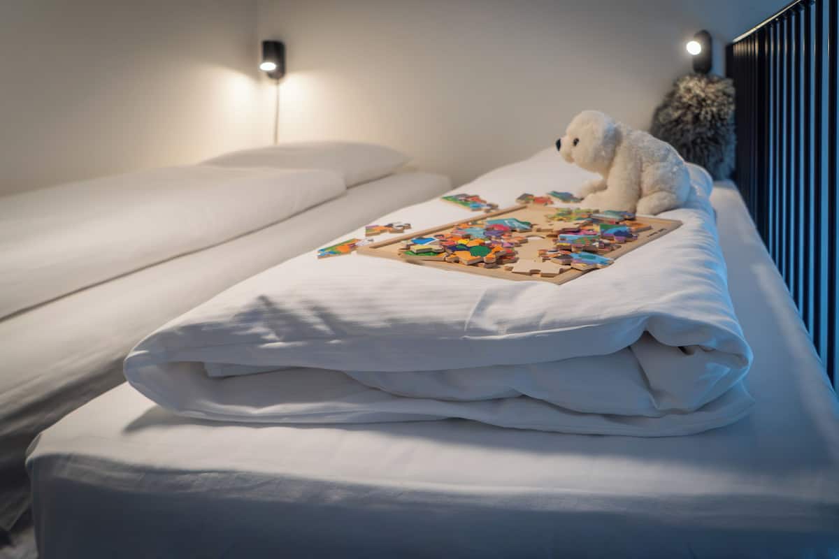 Una cama bien hecha con un rompecabezas encima, un peluche y una lámpara de pared.