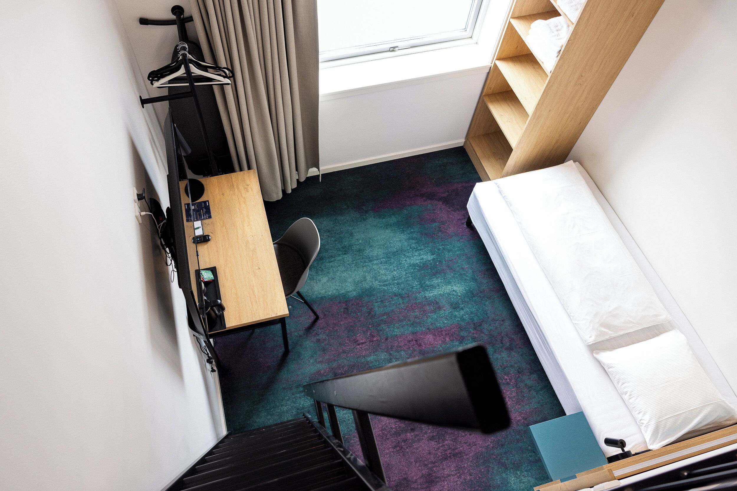 A compact hotel room with a bed, a desk, a chair, a small table, a coat rack, and a window with curtains.