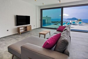 A modern living room with a large flat-screen TV, a sofa with cushions, a small wooden table, and a large sliding glass door leading to an outdoor area with a pool and lounge chairs. - 4 Bedroom Villa with a private pool and incredible views (Peksimet)