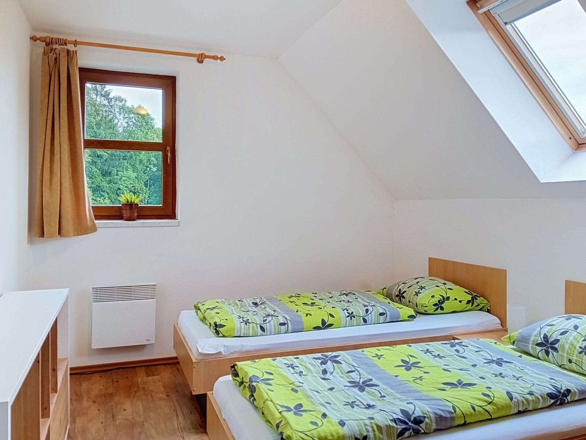 A small, well-lit room with two beds, a window with curtains, a white desk, and a ceiling fan.