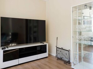 Un salon moderne avec un grand téléviseur à écran plat posé sur un meuble de télévision blanc, une vitrine en verre contenant des verres à vin et un présentoir métallique décoratif.