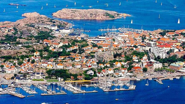 Vue aérienne d’une ville côtière avec un port de plaisance rempli de bateaux, entourée d’un paysage rocheux et de bâtiments résidentiels.