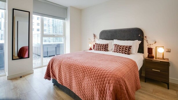 Un dormitorio moderno con una cama grande, una mesita de noche con lámpara y una ventana con vistas a edificios.