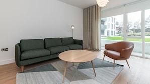 Una sala moderna con sofá, una mesita redonda, un sillón de cuero, una lámpara de pie y ventanas amplias con vistas a edificios y zonas verdes.