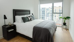 Ein modernes Schlafzimmer mit einem großen Bett, einem Nachttisch, einem Topfpflanzen und Blick auf Gebäude durch ein großes Fenster.