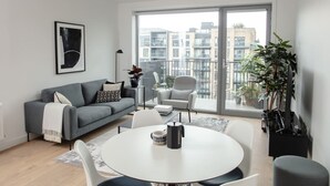 Ein modernes Wohnzimmer mit einem grauen Sofa, weißen Sesseln, einem runden Tisch und einem großen Fenster mit Blick auf die Stadt.