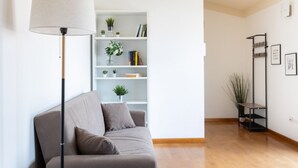 A modern living room with a sofa, a floor lamp, a bookshelf, and a small plant. - Attico Eleonora – Terrazza & Parcheggio in Centro (Oristano)